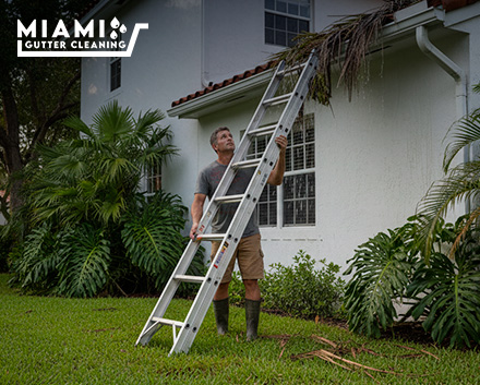 A homeowner looking nervously up at a high, clogged two-story gutter while holding an unstable extension ladder on soft lawn soil.